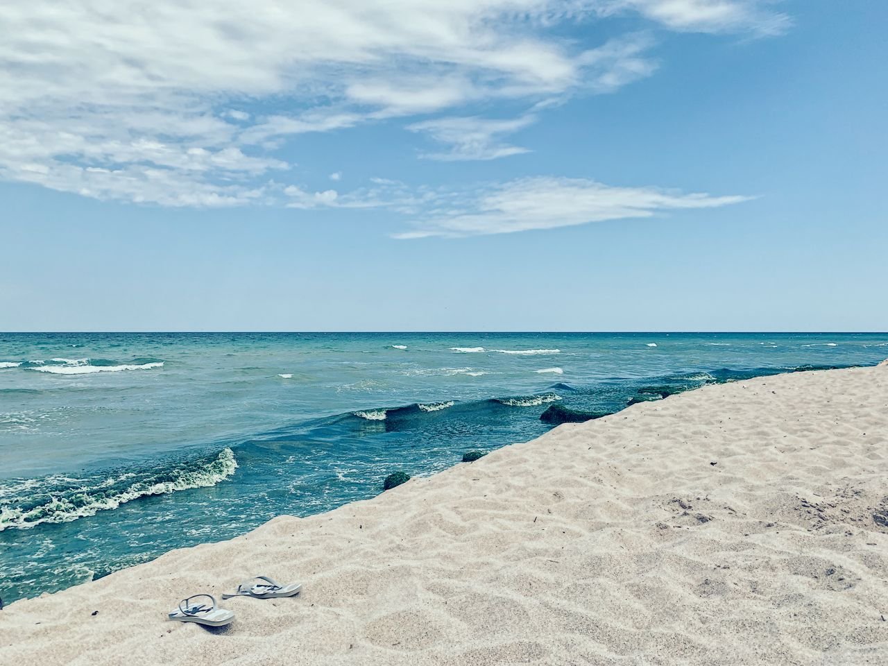 our-mission Small flip flops on sandy shore of vibrant dark blue wavy ocean under sky covered with clouds