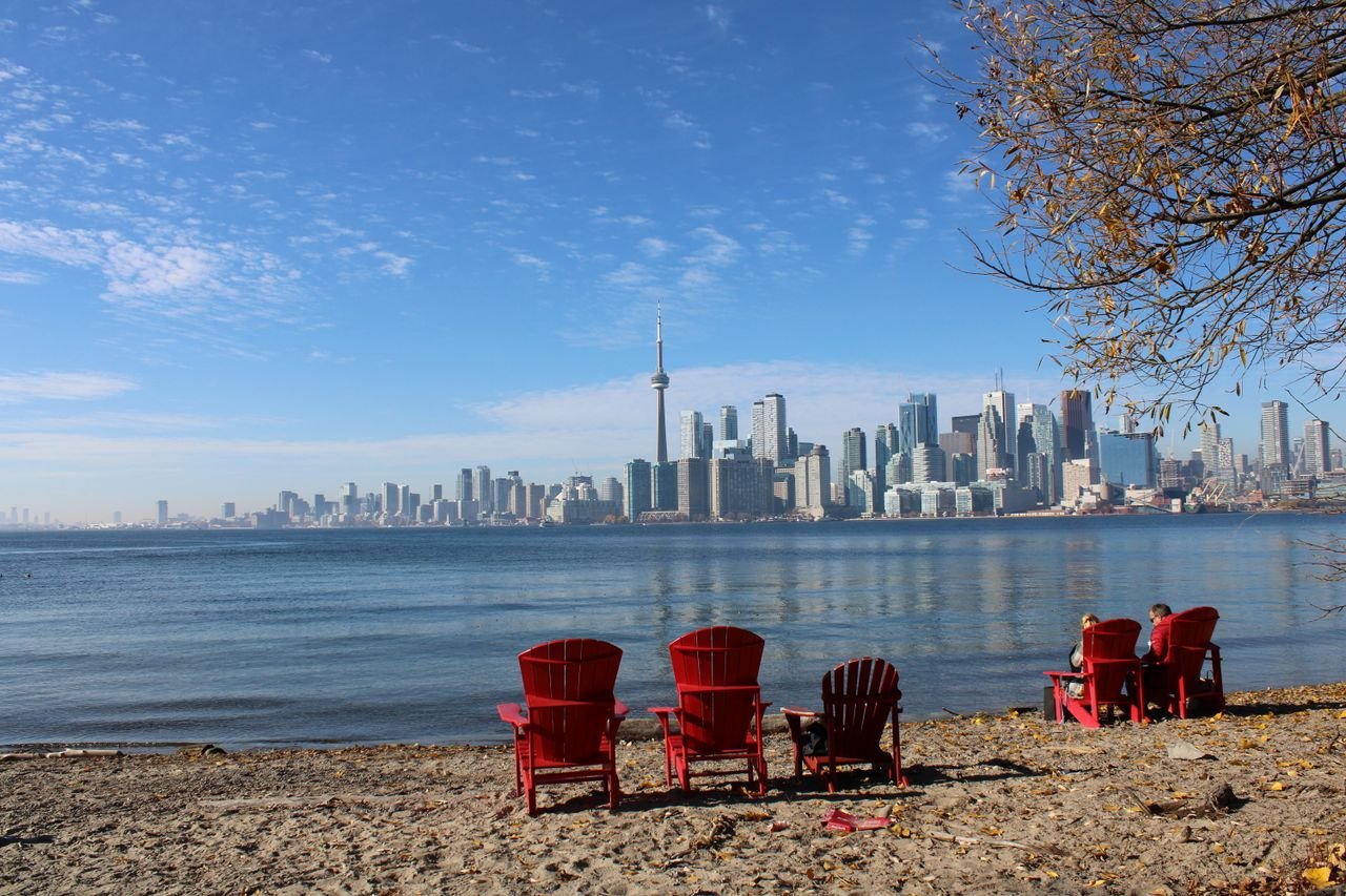 who-we-are Beautiful view of Torontos skyline across the lake with red chairs on a sandy beach.